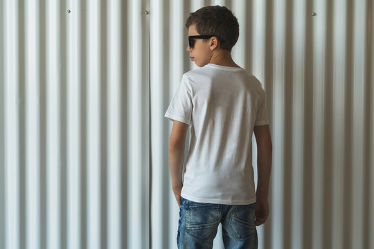 Back view. Young hipster boy dressed in white t-shirt is stands indoor against white wall. Mock up. Space for logo, text, image.