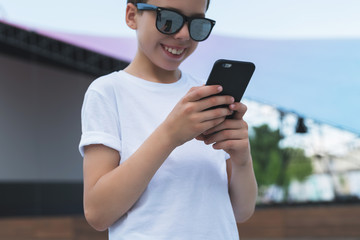 Summer day. Smiling boy in white T-shirt and sunglasses stands outdoor and uses smartphone. Boy plays computer games on digital gadget, surfing internet. Social networks, computer games.