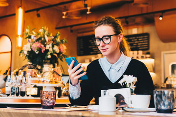 Young business woman in glasses sits in cafe at table, uses smartphone. On table is cup of coffee. Girl working, chatting, blogging, checking email. Online marketing, education, social media, network.