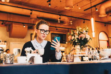 Young business woman in glasses sits in cafe at table, uses smartphone, drinking coffee. Girl working, chatting, blogging, checking email. Online marketing, education, social media, network