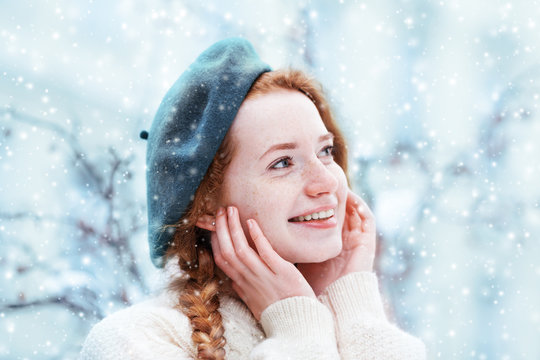 Happy smiling redhead girl with freckles touching her face, looking up. Model posing in street, wearing beret and sweater. Snowfall. Winter, Christmas holidays concept