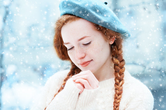 Close up portrait of young beautiful redhead girl with freckles warming her hands, closed eyes. Model posing in street, wearing beret and sweater. Snowfall. Winter, Christmas holidays concept