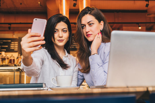 Two young women sit in a cafe at a table in front of a laptop and do selfie on a smartphone. Meeting two friends, colleagues have lunch in a cafe, break during work. Girls blogging, working, learning.