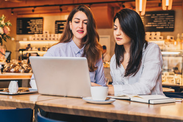 Two young businesswomen sitting in cafe at table and using laptop, working, blogging. Girls are looking at monitor and smiling. Students studying. On desk paper notebook. E-learning, online marketing.