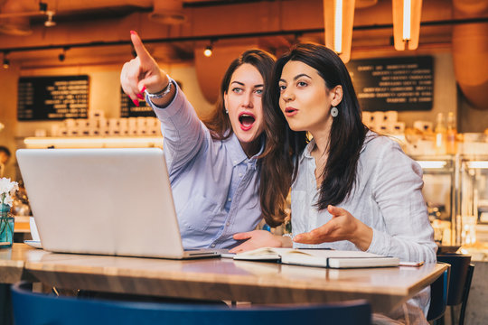 Two young business women are sitting in a cafe at a table in front of a laptop. Girls with joyful surprise point fingers at what they see outside the window. Girls look surprised, delighted, joyful.