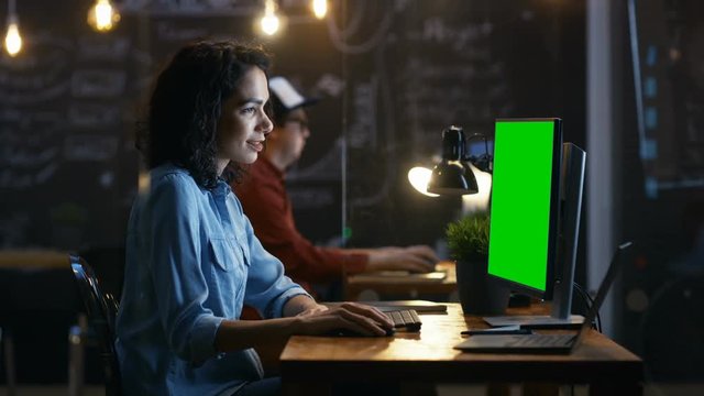 Beautiful Female Office Employee at Her Desktop Works on a Mock-up Green Screen Personal Computer. Her Colleague Sits Beside. Creative Office Evening. RED EPIC-W 8K Camera.