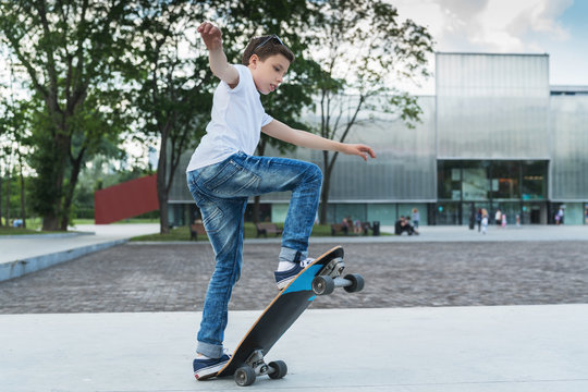 Summer day. The boy is a teenager dressed in a white T-shirt and jeans, skating, doing tricks. In the background is a modern glass building. Vacation, recreation, entertainment, activity.