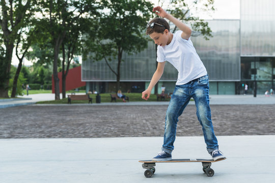 Summer day. The boy is a teenager dressed in a white T-shirt and jeans, skating, doing tricks. In the background is a modern glass building. Vacation, recreation, entertainment, activity.