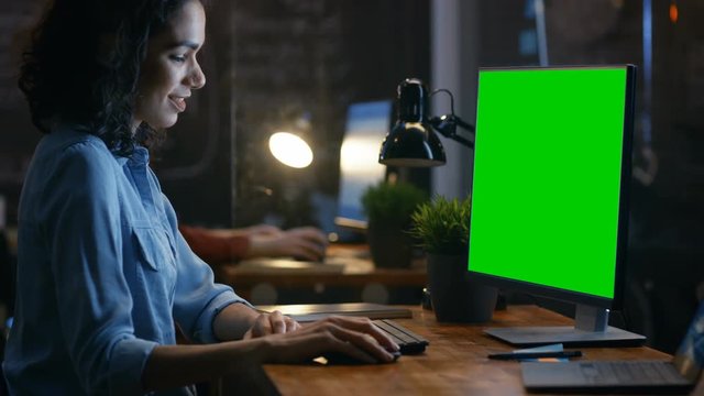 Beautiful Female Office Employee at Her Desk Works on a Mock-up Green Screen Personal Computer. Her Colleague Sits Beside. Creative Office Evening. RED EPIC-W 8K Camera.