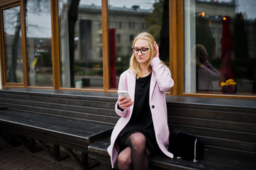 Blonde girl at glasses and pink coat, black tunic sitting on bench at street and looking on smartphone.