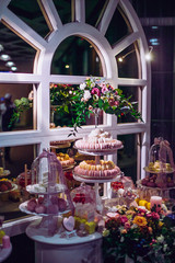 White wedding cake decorated by flowers standing on festive table with lots of snacks on side.