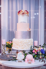 White wedding cake decorated by flowers standing on festive table with lots of snacks on side.