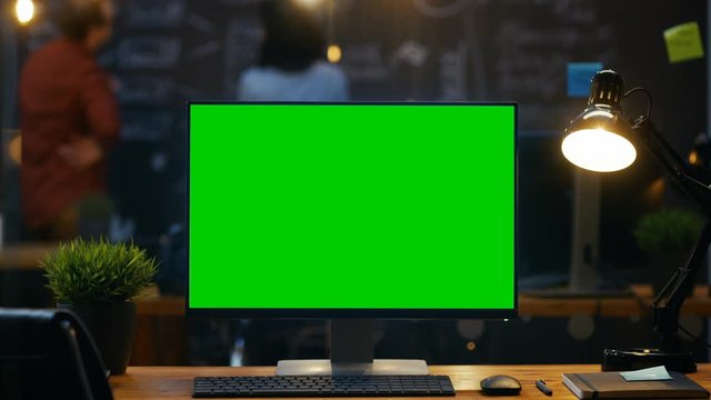 Personal Computer with Mock-up Green Screen Monitor Stands on the Office Desk, In the Background People Work on a Blackboard Wall in the Creative Office. RED EPIC-W 8K Camera.