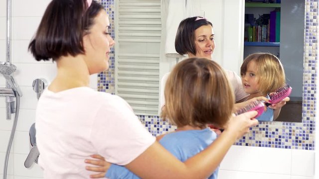 Happy mother hugging little daughter in front of mirror brushing blond hair
