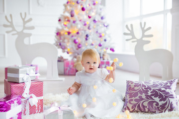 Fototapeta premium Beautiful little baby celebrates Christmas. New Year's holidays. Sweet baby girl in cute dress having fun in festive decorated room.