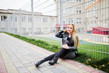 Blonde fashionable girl in long black leather coat against iron fence at street.