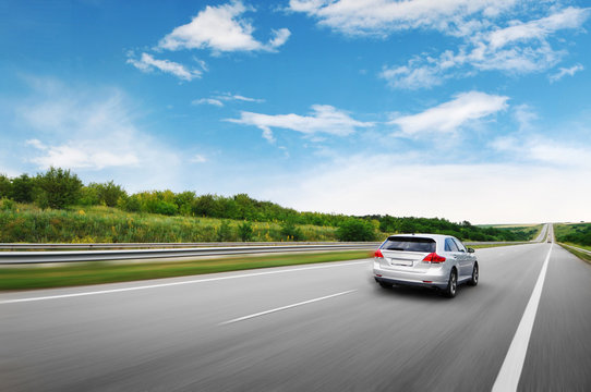 A Silver Car Driving Fast On The Countryside Road Against Sky With Clouds