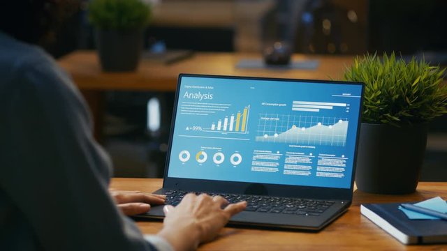 Female Analyst at Her Desk Works on a Laptop Showing Statistics, Graphs and Charts. She Works on the Wooden Table in Creative Office. Over the Shoulder Footage. RED EPIC-W 8K Camera.