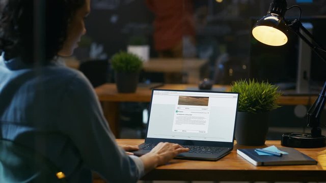 Female Uses Laptop for Browsing Social Networks Site, Scrolling Webpage Wall. Over the Shoulder Footage. In the Background in this Creative Office. 