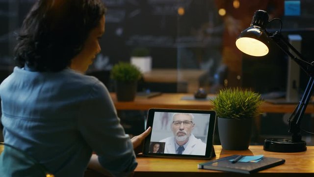 Beautiful Woman Sits at Her Desk and Holds Tablet Computer, She Talks with Her Doctor Via Video Call. Creative Environment in the Evening Office. RED EPIC-W 8K Camera.