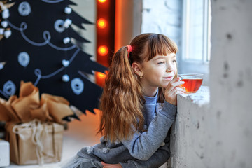 Beautiful child and drinks tea and looks out the window. Concept Happy Christmas, New Year, holiday, winter, childhood.