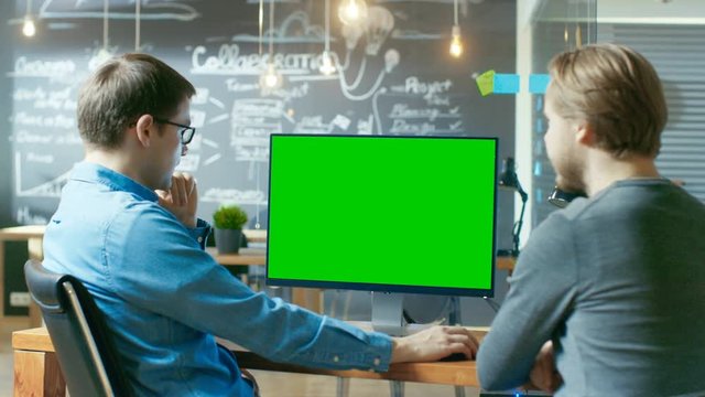 Two Office Employees Have Project Related Discussion, on the Desk Stands Personal Computer with Mock-up Green Screen. They Work in the Creative Office Environment. RED EPIC-W 8K Camera.
