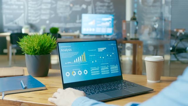 Male Analyst at His Desk Works on a Laptop Showing Statistics, Graphs and Charts. He Works in Light and Modern Creative Office.RED EPIC-W 8K Camera.