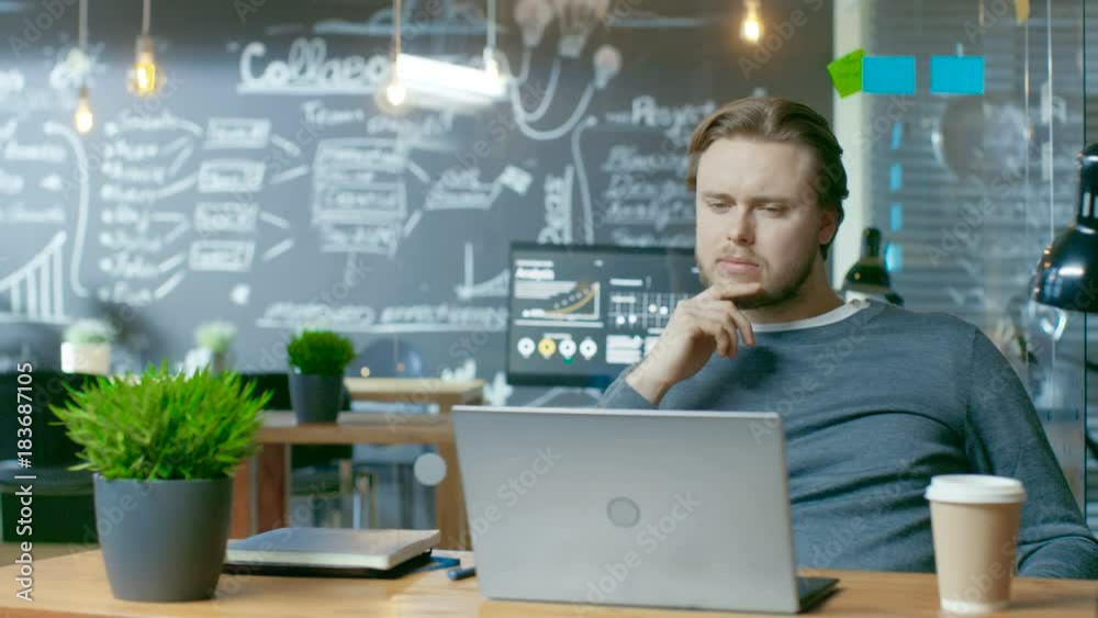 Poster Handsome Young Office Employee Thinks on a Problem Solution While Working on a Laptop Computer. He's Working in the Creative Stylish Office. RED EPIC-W 8K Camera.