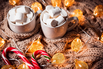 Christmas background with cocoa or coffee with marshmallows and candy in a white cup on a brown knitted winter scarf and a glowing golden garland. Beautiful concept of home comfort and warmth