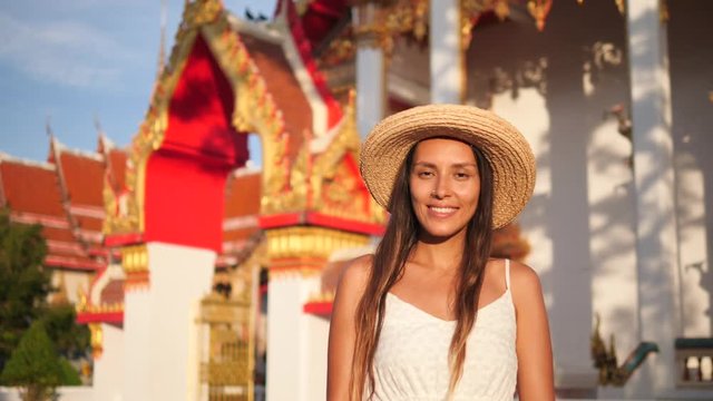 Beautiful Smiling Young Mixed Race Tourist Woman in Straw Hat with Traditional Thai Buddhist Temple on Background. Thailand. 4K, Slowmotion.