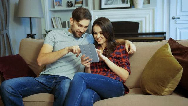 Portrait of young attractive couple sitting on the couch, scrolling and taping on the tablet device in the cozy and nice room at home. Indoor