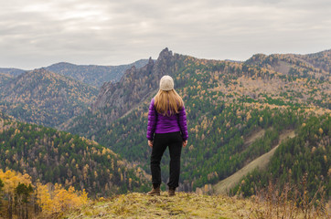 A girl in a lilac jacket standing on a mountain, a view of the mountains and an autumn forest by a cloudy day