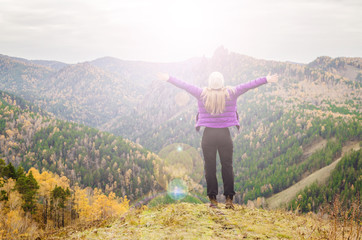 Fototapeta na wymiar A girl in a lilac jacket stretching her arms on a mountain, a view of the mountains and an autumn forest by a cloudy day, free space for text