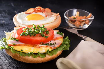 Breakfast. Fried egg on a roll, tomato, greens, bacon, mushrooms. Cutlery. Close-up Black background