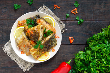 Pieces of roasted carp with lemon and greens on a plate on a dark wooden background. Top view.