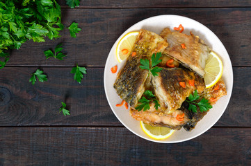 Pieces of roasted carp with lemon and greens on a plate on a dark wooden background. Top view.