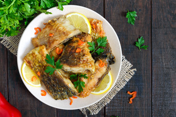 Pieces of roasted carp with lemon and greens on a plate on a dark wooden background. Top view.