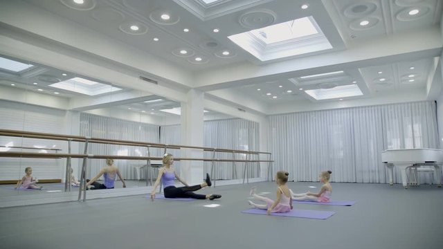 The young ballet teacher shows how to point toes with straight legs to two girls. The lady sit in front of the little girls on the mat and raise her feet one by one and the little dancers are