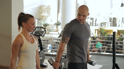 Young couple doing biceps dumbbells curls in gym indoors. Man and woman stand and raise free weight to shoulder level, bending arms in elbows, looking at each other with cheerful smiles. Beautiful - Powered by Adobe