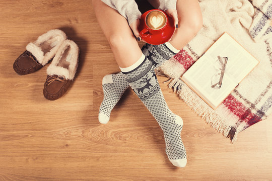  Young woman drinking cappuccino and sitting on the wooden floor. Close-up of female legs in warm socks with a deer, book and coffee vintage toned image, top view. Comfort winter holidays concept 