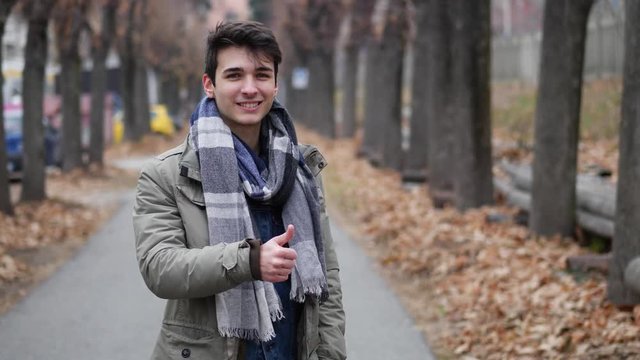 Handsome young man among trees, looking at camera doing thumb up sign for OK,, wearing a winter coat, looking at camera with a smile