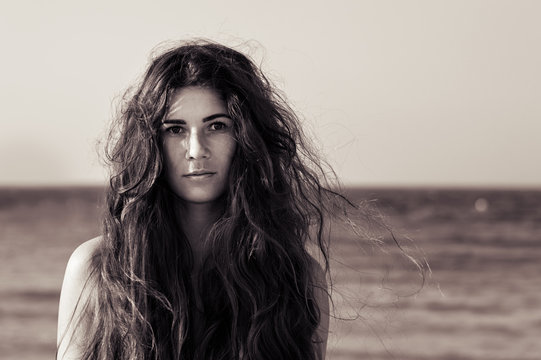 Portrait of female model with messy long hair standing at the seaside. Black and white photo of young woman with confident look