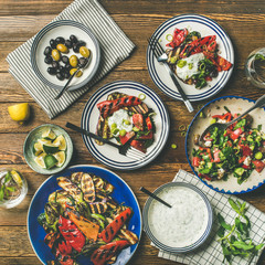 Flat-lay of healthy dinner table setting. Fresh salad, grilled vegetables with yogurt sauce, pickled olives, lemon water over wooden background, top view, square crop. Clean eating, vegetarian food