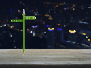 2018 and 2017 direction sign plate with green pencil on wooden table over blur colorful night light of modern city tower, Business happy new year planning concept