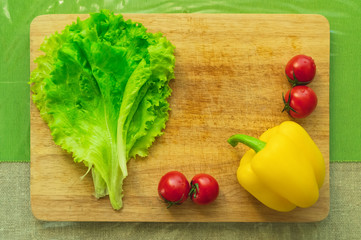 Fresh green lettuce leaves lie on a wooden cutting board next to the yellow bell pepper and red cherry tomatoes on a green table