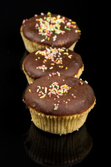 Vanilla cupcakes with chocolate cream and colorful sprinkles above black reflections background