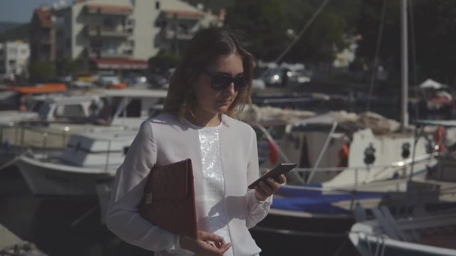 business woman with smartphone in formal clothes standing on the dock against background of blurry boats and prints a message