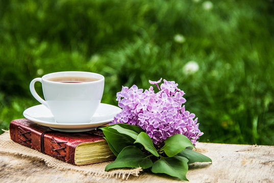 Cup of hot tea, book and branch of lilac. Tea in the spring garden. Home cosiness. Copy space
