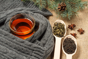 Obraz na płótnie Canvas Cup of tea with slice of lemon wrapped up in wool scarf, wooden spoons with leaves of green and black tea, natural fir tree branches with cone
