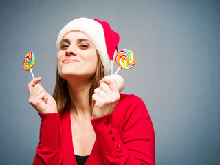 Fototapeta premium Cheerful young girl in Santa's hat with bright lollipops in hands on a blue background. Celebration of new year and Christmas